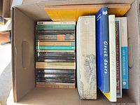 Box with mix of paperbacks and hardcovers; visible art monographs "De Chirico", "Greek Doors"