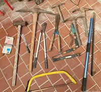 Floor layout of assorted long and hand tools including mattocks, saws, loppers and sledgehammer