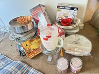 Group shot of boxed hand mixer, boxed can opener, stack of metal cake pans, copper Bundt pan, rolling pin, measuring cup, heart ramekin, decorating set box, glass jar, pink candles