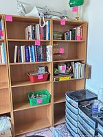 Two tall light oak veneer bookcases filled with books and a black plastic drawer tower beside them