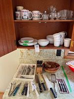Overview of cabinet and counter showing Corelle dishes, mugs, flatware, knives, glassware