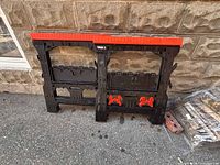 Front view of both folded sawhorses leaning against wall showing red tops and orange brackets