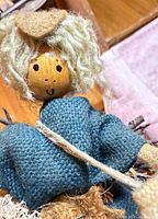 Front view of doll showing face, burlap hat, raffia hair, twig bundle and blue cloth outfit