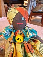 Front view of cloth rag doll showing floral dress, head wrap, bead earrings and face details with yellow sticker