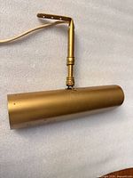 Gold brass-tone cylindrical picture light laying flat, showing adjustable arm and cord