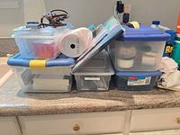 Top view of stacked clear storage bins with blue lids, smaller bin on top, ribbon spool, cables