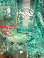 Two promotional glasses—Grapette logo in white, Monette logo in red—resting on bubble wrap