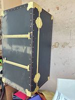 Full view of closed trunk showing dark covering, brass hardware, corner protectors, side straps
