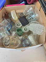 Top view of assorted glassware in box showing gold lattice tumbler, blue-tinted glasses, green dessert cups, pressed glass pieces and pouches