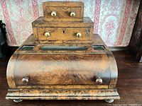 Front view of burl walnut stationery box showing stepped drawers, curved front panel, brass knobs and bun feet