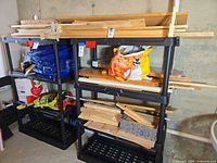 Two black plastic four-tier shelving units with contents resting on them (contents not included) against a basement wall.