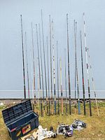 Group of 18 upright rods beside blue tackle box and several reels