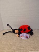 Full view of Ann Geddes Baby Ladybug plush doll lying on side with hang tag visible