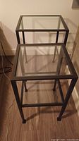 Two black metal frame side tables with clear glass tops viewed from front angle