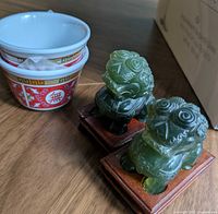 Two green stone guardian lion figurines on wood stands beside bowls