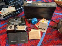 Overall shot showing Polaroid camera in case, Minolta camera, flashgun, blue bulb, GE M3 box, manuals, and carrying case