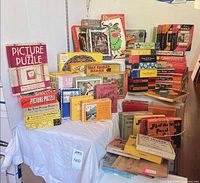 Overall lot showing stacks of 30–40 vintage jigsaw puzzle boxes on table