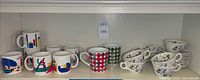 Shelf view of entire lot; novelty print mugs, polka-dot mugs, floral teacups and saucers visible