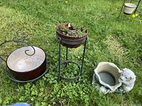 Three plant stands on grass: wrought iron with glass bowl, wrought iron column with metal pot, ceramic bear planter