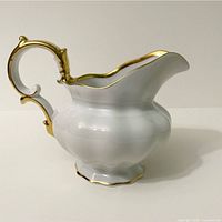 Full left side view of white porcelain pitcher with gold trim and ornate handle