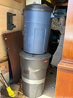 Two stacked Rubbermaid garbage cans; blue can on top, dark gray can with duct tape repair on bottom, lids visible