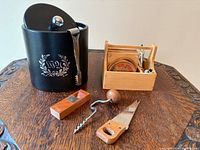 All items grouped on tabletop: black faux leather ice bucket with lid and tongs, wooden caddy with bar tools and coasters, separate corkscrew and saw knife visible