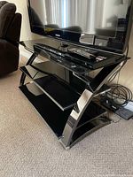 Front angled view of black glass three-shelf TV stand showing curved chrome frame and open shelving