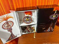 Open box showing Barbie doll with feathered headdress, patterned gown and stand inside packaging, plus certificate artwork and outer box