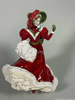 Front three-quarter view of porcelain figurine showing red gown, white fur muff and trim, green bonnet