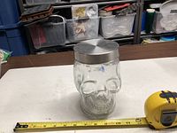 Front view of clear glass skull jar with metal lid beside tape measure for scale