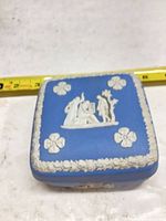 Top view showing blue box with white relief scene and floral border, tape for scale