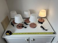 Five dusty pink rechargeable lamps with cords on white cabinet, one lamp lit, measuring tape for scale