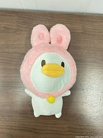 Front view of plush showing duck face, pink bunny hood, yellow beak and chest bell