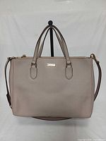 Front view of taupe Kate Spade satchel on stand showing logo, handles and attached strap