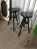 Two black adjustable bar stools with round seats and metal footrests standing on a floor inside a metal storage unit.
