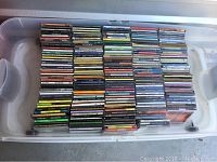 View of the mixed CDs stacked in a clear plastic bin, showing many CD jewel cases with visible wear and varied colors of spines.