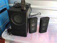 Full view of subwoofer, two satellite speakers and wired remote control on plastic table.