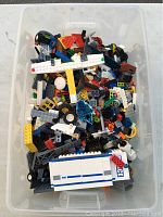 Plastic bin filled with assorted LEGO pieces in various colors and shapes including wheels and panels.