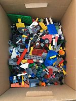 Top-down view of a box filled with assorted LEGO bricks including basic plates, wheels, and stones in different colors.