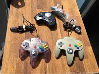 Photo showing two Nintendo 64 controllers (one translucent purple, one beige) and one black Venturer wired controller on a wooden surface.