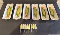Six ceramic corn motif trays and eight knives laid out