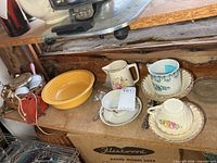 Shelf view showing yellow Fiesta bowl, small pitcher, teacup with saucer, cream bowls in wicker holders
