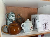 Group shot of all items on shelf: enamel kettle, brown teapot, wire rack, white floral teapot, boxed fondue set