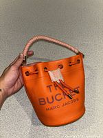 Front view of orange Marc Jacobs bucket bag showing drawstring, striped handle and front logo text