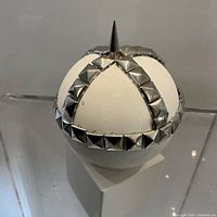 Front angled view of spherical sculpture on plinth showing metal studded cross pattern and spike