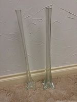 Full view of both clear glass trumpet bud vases standing upright on carpet against wall
