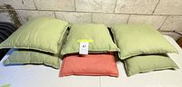 Stack of six square outdoor throw pillows, five light green and one coral, on table