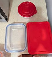 Round plastic container with red lid next to two rectangular glass containers, one with red lid, one base only