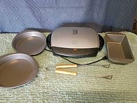 Closed George Foreman grill flanked by two round pans, loaf pan, and two skewers