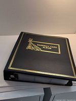 Closed black binder showing gold foil "Baseball Card Album" cover